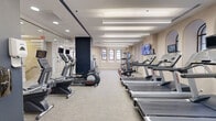This fitness studio offers a thoughtfully designed single-floor exercise space that combines architectural charm with a comprehensive selection of modern workout equipment. The room is characterized by its elongated layout, neutral-toned walls, and light gray carpet flooring, creating a clean and focused environment for exercise.

Architecturally, the studio stands out with a series of elegant arched windows fitted with black-framed grilles, allowing natural light to filter into the space and lending the interior a distinctive, refined character. Linear pendant lighting runs the length of the ceiling, ensuring consistent, bright illumination throughout.

The exercise room is outfitted with a wide variety of cardio equipment, including multiple treadmills, elliptical machines, stair climbers, and stationary and spin bikes. A dedicated free weights area features a dumbbell rack for strength training, while additional cable and resistance machines provide options for full-body workouts. Wall-mounted flat-screen TVs are positioned throughout the room, offering entertainment during workouts.

A full-length mirror along one wall visually expands the space and supports form-focused training. A bold navy blue accent wall adds a striking visual contrast to the otherwise neutral palette. The studio's layout ensures ample circulation space between equipment, contributing to a comfortable and functional workout environment.