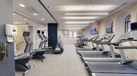 This fitness studio offers a thoughtfully designed single-floor exercise space that combines architectural charm with a comprehensive selection of modern workout equipment. The room is characterized by its elongated layout, neutral-toned walls, and light gray carpet flooring, creating a clean and focused environment for exercise.

Architecturally, the studio stands out with a series of elegant arched windows fitted with black-framed grilles, allowing natural light to filter into the space and lending the interior a distinctive, refined character. Linear pendant lighting runs the length of the ceiling, ensuring consistent, bright illumination throughout.

The exercise room is outfitted with a wide variety of cardio equipment, including multiple treadmills, elliptical machines, stair climbers, and stationary and spin bikes. A dedicated free weights area features a dumbbell rack for strength training, while additional cable and resistance machines provide options for full-body workouts. Wall-mounted flat-screen TVs are positioned throughout the room, offering entertainment during workouts.

A full-length mirror along one wall visually expands the space and supports form-focused training. A bold navy blue accent wall adds a striking visual contrast to the otherwise neutral palette. The studio's layout ensures ample circulation space between equipment, contributing to a comfortable and functional workout environment.