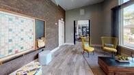 This thoughtfully designed single-floor amenity space offers a dynamic blend of industrial and contemporary aesthetics. Wood-look tile flooring runs throughout, complementing the warm, neutral color palette of the sage-gray walls and crisp white trim. Soaring ceilings amplify the sense of openness across the generously proportioned layout.

The lounge area features plush light-blue seating arranged around a circular coffee table, with dramatic floor-to-ceiling dark drape panels framing large windows that look out onto a resort-style pool and lush outdoor surroundings, flooding the space with natural light. Adjacent to the lounge, a game and social zone showcases a striking exposed brick accent wall, a wall-mounted television, and a communal high-top table with bar stools — ideal for casual gatherings.

A dedicated games corner offers a large Scrabble board, chalkboard wall, and eclectic seating with vibrant yellow accent chairs and colorful ottomans, injecting a playful energy into the space. A built-in workstation with a desktop computer and pendant lighting provides a functional nook for productivity. A textured 3D-panel accent wall and a beverage refrigerator add further distinctive touches. The open floor plan connects each area seamlessly, creating a versatile, inviting environment suited for both relaxation and community engagement.