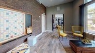 This thoughtfully designed single-floor amenity space offers a dynamic blend of industrial and contemporary aesthetics. Wood-look tile flooring runs throughout, complementing the warm, neutral color palette of the sage-gray walls and crisp white trim. Soaring ceilings amplify the sense of openness across the generously proportioned layout.

The lounge area features plush light-blue seating arranged around a circular coffee table, with dramatic floor-to-ceiling dark drape panels framing large windows that look out onto a resort-style pool and lush outdoor surroundings, flooding the space with natural light. Adjacent to the lounge, a game and social zone showcases a striking exposed brick accent wall, a wall-mounted television, and a communal high-top table with bar stools — ideal for casual gatherings.

A dedicated games corner offers a large Scrabble board, chalkboard wall, and eclectic seating with vibrant yellow accent chairs and colorful ottomans, injecting a playful energy into the space. A built-in workstation with a desktop computer and pendant lighting provides a functional nook for productivity. A textured 3D-panel accent wall and a beverage refrigerator add further distinctive touches. The open floor plan connects each area seamlessly, creating a versatile, inviting environment suited for both relaxation and community engagement.