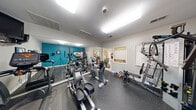 This fitness center is a thoughtfully outfitted exercise space designed to meet a variety of workout needs within a compact footprint. The room features a bold teal accent wall that adds energy and visual interest, contrasting with the neutral gray walls and bright, flush-mounted ceiling lighting that keeps the space well-illuminated throughout.

The flooring is durable rubber gym matting, ideal for both cardio and strength training activities. A large wall-spanning mirror along one side visually expands the room and assists users in monitoring their form during exercise. The cardio equipment lineup includes elliptical trainers, upright and recumbent stationary bikes, and a TRUE-brand treadmill, offering a well-rounded selection for aerobic workouts. Strength training is supported by a multi-station cable machine, a weight bench, and an assortment of free weights and dumbbells.

A wall-mounted television provides entertainment during workouts, while motivational framed fitness photography decorates the walls. The space includes a door with a glass panel that allows natural light to enter and provides a view to the outdoors. An adjacent interior window and connecting door offer access to a neighboring room. A fire extinguisher is also visibly mounted for safety. Overall, the fitness center delivers a functional and motivating environment for daily exercise.