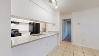 This single-floor unit offers a practical and functional layout designed for comfortable everyday living. The open-concept living and kitchen area is finished with large neutral-toned ceramic floor tiles that extend throughout the common spaces, creating a bright and cohesive feel. The kitchen features a peninsula-style counter with a double stainless steel sink, black appliances including a refrigerator and a coil-top electric range with overhead ventilation hood, white cabinetry, and track lighting above. A dedicated in-unit laundry area is conveniently situated just off the kitchen. The living area is spacious and open, benefiting from a clean, minimalist design with white walls and a textured ceiling. A separate room with large sliding glass doors fitted with vertical blinds allows natural light to filter in and provides outdoor views, making it a versatile space suitable as a living room or bedroom. Additional rooms feature plush carpeting in neutral gray tones, offering a softer, quieter environment. The bathroom is clean and well-appointed, featuring a white vanity with under-sink storage, a wall mirror, tile-surround bathtub and shower combination, and light tile flooring. The unit also includes a closet with bi-fold doors, providing ample storage throughout.