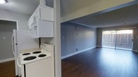 This single-floor space is designed with a practical and clean aesthetic throughout. The galley-style kitchen is outfitted with white cabinetry providing ample storage, a stainless steel sink set into light-toned countertops, and a full-size white refrigerator. A ceiling fan with a light fixture overhead adds both function and airflow to the space. The kitchen features wood-look flooring that carries a warm tone, complementing the neutral gray walls and crisp white trim. A textured ceiling runs throughout the unit, adding subtle visual interest.

Adjacent to the kitchen, a stacked washer and dryer unit offers in-unit laundry convenience. The electric range nearby keeps all major appliances centrally accessible. The kitchen opens directly into the main living area, which is notably open and airy. Dark hardwood-style flooring anchors the living space, creating a rich contrast against the soft gray walls and white baseboards. A sliding glass door fitted with vertical blinds allows natural light to filter into the room while providing access to an outdoor area. The overall ambiance is neutral, clean, and move-in ready.