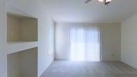This single-story space features a clean, functional layout designed to maximize comfort and usability. The main living area is finished with soft, wall-to-wall carpeting that adds warmth underfoot, while crisp white walls and smooth ceilings create a bright, airy atmosphere throughout. A ceiling fan is positioned overhead, providing efficient air circulation and supplemental lighting to the room.

Natural light is a defining characteristic of this space, courtesy of a generously sized sliding glass door fitted with vertical blinds. These blinds offer flexible light control, allowing occupants to easily adjust brightness levels and privacy as needed. The sliding door also provides convenient access to an outdoor area.

Adding to the room's practicality, built-in shelving is recessed into one wall, offering dedicated storage or display space without encroaching on the floor area. A framed window on the adjacent wall introduces additional natural light, further enhancing the open, welcoming feel of the interior.

Overall, the space presents a neutral, move-in-ready environment with thoughtful built-in features, ample natural light, and a straightforward single-floor layout well-suited for everyday living.