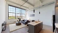 This single-floor industrial loft blends raw architectural character with modern finishes throughout. The open-concept main living area is anchored by a dramatic wall of floor-to-ceiling factory-style steel-framed windows that flood the space with natural light and frame sweeping outdoor views. Exposed ductwork and white-painted wood beam ceilings soar overhead, reinforcing the loft's industrial heritage while maintaining a bright, airy atmosphere. Polished concrete floors run throughout, tying the space together seamlessly.

The kitchen is a standout feature, equipped with stainless steel appliances including a refrigerator, range, and microwave, complemented by subway tile backsplash, two-tone cabinetry in white and warm gray, and a generous island with a quartz countertop and bar seating beneath sleek pendant lighting. The living and dining areas flow naturally from the kitchen, creating an ideal layout for entertaining.

A hallway with a sliding barn door leads to the private bedroom, which enjoys tall ceilings and ample natural light filtered through high windows. A dedicated utility area houses the water heater and laundry equipment alongside an electrical panel. The space offers a compelling combination of industrial character, contemporary design, and functional living.