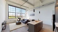 This single-floor industrial loft blends raw architectural character with modern finishes throughout. The open-concept main living area is anchored by a dramatic wall of floor-to-ceiling factory-style steel-framed windows that flood the space with natural light and frame sweeping outdoor views. Exposed ductwork and white-painted wood beam ceilings soar overhead, reinforcing the loft's industrial heritage while maintaining a bright, airy atmosphere. Polished concrete floors run throughout, tying the space together seamlessly.

The kitchen is a standout feature, equipped with stainless steel appliances including a refrigerator, range, and microwave, complemented by subway tile backsplash, two-tone cabinetry in white and warm gray, and a generous island with a quartz countertop and bar seating beneath sleek pendant lighting. The living and dining areas flow naturally from the kitchen, creating an ideal layout for entertaining.

A hallway with a sliding barn door leads to the private bedroom, which enjoys tall ceilings and ample natural light filtered through high windows. A dedicated utility area houses the water heater and laundry equipment alongside an electrical panel. The space offers a compelling combination of industrial character, contemporary design, and functional living.
