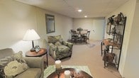 This multi-story space spans three floors, offering a well-rounded layout designed to accommodate a variety of living and functional needs. The finished lower level is a standout feature, providing a cozy and inviting atmosphere with a drop-tile ceiling and recessed lighting that creates a warm, evenly lit environment. This level is thoughtfully arranged to serve as both a lounging and dining area, with plush, patterned armchairs and a sofa grouped around a coffee table on a decorative area rug, alongside a bistro-style dining set with wrought-iron chairs positioned toward the rear of the space. A framed mirror on the wall adds depth and brightness to the room, while a side console table with decorative accents contributes to the finished, homey aesthetic. The neutral carpeting throughout the lower level ties the space together with a clean, understated look. Accent lighting from a classic table lamp enhances the comfortable ambiance. With its generous open floor plan on this level and two additional floors above, the overall space provides ample room for both private retreats and shared social gatherings, making it a functional and welcoming environment throughout.