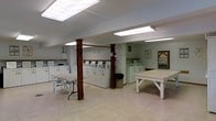 This single-floor communal laundry facility is designed with practicality and convenience in mind. The space is generously sized, featuring multiple top-load washing machines and dryers arranged in rows along the walls, providing ample capacity for residents. Overhead fluorescent lighting illuminates the entire area evenly, complemented by what appears to be a skylight panel in the ceiling that adds a touch of natural brightness to the environment. The flooring consists of durable light-colored tile, well-suited for a high-traffic utility space. Sturdy wooden support columns, finished in a warm reddish-brown tone, punctuate the room and add a subtle architectural character. Several large white folding tables are positioned throughout the space, offering convenient surfaces for sorting and folding laundry. A waste bin is centrally located for easy access. The walls are kept clean and neutral, with a bulletin board area near the entrance featuring posted notices and informational materials. The overall layout is open and uncluttered, ensuring easy movement throughout the facility. This space is thoughtfully organized to serve a multi-unit residential community efficiently.