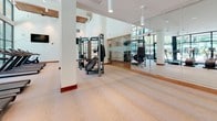 This fitness center is designed with both functionality and aesthetics in mind, offering a bright, open environment suited for a comprehensive workout experience. The space is anchored by expansive floor-to-ceiling glass walls that frame views of palm trees and an outdoor courtyard, flooding the interior with natural light and creating an airy, energizing atmosphere.

The main workout floor features light wood-style flooring and soaring ceilings supported by structural columns, contributing to the generous sense of scale throughout. Full-length mirrored walls along one side visually expand the space while allowing users to monitor their form. The cardio zone is well-appointed with multiple treadmills, ellipticals, and stationary bikes arranged to take advantage of the outdoor views. A dedicated strength and functional training area includes multi-station cable machines and free-standing equipment from recognized brands.

An adjoining reception or check-in area connects to the fitness floor, featuring warm wood cabinetry, soft carpeted flooring, and a glass-paneled entryway with an elegant gold-trimmed door. This transitional space adds a refined, hospitality-inspired touch to the overall facility.