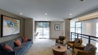 This single-floor space offers a thoughtfully designed interior that blends comfort and functionality. The main lounge area is anchored by a plush blue-gray sofa accented with warm terracotta pillows, complemented by a pair of mustard-yellow armchairs arranged around a sculptural round wooden coffee table. Recessed lighting in the smooth white ceiling provides consistent illumination throughout the space, while sheer curtains allow abundant natural light to filter through large windows that frame views of lush greenery outside. The neutral wall tones in soft taupe and white create a calm, inviting atmosphere. A dedicated workspace is visible in the background, featuring a sleek desk and task chair positioned near a window, maximizing natural light for productivity. Abstract wall art adds a refined artistic touch to the space. The light-toned carpeting ties the seating areas together seamlessly. A staircase with a modern railing is partially visible, suggesting connectivity to additional levels. Overall, the space balances a professional yet welcoming ambiance.