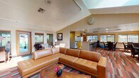 This single-floor space showcases a thoughtfully designed open-concept layout that seamlessly connects the living, dining, and kitchen areas. The living room serves as the central hub, anchored by a large caramel-toned leather sectional sofa positioned atop a multi-colored striped area rug. Vaulted ceilings with track lighting elevate the sense of openness, while a skylight introduces additional natural light overhead. A flat-screen television and media console occupy one wall, complemented by a cozy seating nook featuring a fireplace framed by dark accent walls and artwork above the mantle — creating a distinct, intimate atmosphere within the larger open plan.

The kitchen is well-equipped with warm honey-toned cabinetry, white appliances, and a central island with bar seating. A decorative track light fixture adds character overhead. Adjacent to the kitchen, the dining area accommodates a dark-toned table with upholstered chairs, surrounded by wood-framed windows with horizontal blinds that filter natural light while maintaining privacy. An entry door with exterior visibility rounds out this zone. Throughout the space, multicolored wood-plank flooring ties the areas together, contributing warmth and visual continuity across the entire floor plan.