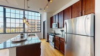 Located at 272 S Front St in the heart of Columbus, Ohio, this single-floor loft-style unit blends industrial character with modern finishes throughout. The open living area is a standout, anchored by warm hardwood floors and floor-to-ceiling steel-framed windows that flood the space with natural light and frame dynamic views of the surrounding downtown cityscape. Exposed ductwork along the ceilings reinforces the industrial aesthetic, while crisp white walls keep the overall ambiance bright and airy.

The kitchen is thoughtfully designed with rich dark wood cabinetry, granite countertops, a subway tile backsplash, and a full suite of stainless steel appliances including a refrigerator, range, and built-in microwave. A central island with pendant and track lighting provides both additional workspace and casual seating, making the kitchen as functional as it is visually appealing.

The space includes two full bathrooms. One features a walk-in glass-enclosed shower with white tile surround, a granite-topped vanity, and tile flooring. The second offers a full soaking tub with a glass sliding door, matching granite vanity, and the same clean tile aesthetic. The bedroom areas benefit from the large steel-framed windows, continuing the loft's open and light-filled character.