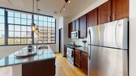 Located at 272 S Front St in the heart of Columbus, Ohio, this single-floor loft-style unit blends industrial character with modern finishes throughout. The open living area is a standout, anchored by warm hardwood floors and floor-to-ceiling steel-framed windows that flood the space with natural light and frame dynamic views of the surrounding downtown cityscape. Exposed ductwork along the ceilings reinforces the industrial aesthetic, while crisp white walls keep the overall ambiance bright and airy.

The kitchen is thoughtfully designed with rich dark wood cabinetry, granite countertops, a subway tile backsplash, and a full suite of stainless steel appliances including a refrigerator, range, and built-in microwave. A central island with pendant and track lighting provides both additional workspace and casual seating, making the kitchen as functional as it is visually appealing.

The space includes two full bathrooms. One features a walk-in glass-enclosed shower with white tile surround, a granite-topped vanity, and tile flooring. The second offers a full soaking tub with a glass sliding door, matching granite vanity, and the same clean tile aesthetic. The bedroom areas benefit from the large steel-framed windows, continuing the loft's open and light-filled character.