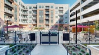 Located at 1345 14th St in Plano, Texas, this outdoor amenity space is set within a modern, multi-story residential community defined by a striking contemporary façade combining brick, metal panel cladding, and large-format windows. The centerpiece of the courtyard is a resort-style pool enclosed by sleek black metal fencing, surrounded by ample deck space furnished with lounge chairs, colorful umbrellas, and stylish outdoor seating arrangements — ideal for relaxation and socializing under the Texas sun. Adjacent to the pool area, a spacious paved courtyard features comfortable outdoor sofas with accent pillows and a low coffee table, creating an inviting lounge setting. A dedicated activity zone includes a full-size ping-pong table positioned beneath a covered, semi-enclosed corridor, which is finished with recessed lighting, large-format ceiling panels, and textured stone paver flooring that extends throughout the outdoor space. Oversized geometric planters with lush tropical foliage add greenery and visual interest throughout. A grand entry point with floor-to-ceiling black steel-framed glass doors provides a refined transition between the interior and exterior, filling the covered passage with natural light. The overall ambiance is modern, polished, and community-oriented, offering a well-designed outdoor environment for residents to gather and unwind.