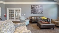 Located at 7799 W Pearson in Shelby Township, Michigan, this two-story space is thoughtfully designed to serve as a welcoming gathering or community environment. The interior is finished in a consistent cool gray palette with teal accent walls, patterned carpeting, and white trim details that lend a clean, contemporary feel throughout.

The main level features a comfortable lounge area furnished with tufted leather seating, upholstered accent chairs in coordinating patterns, and a central coffee table. Large windows fitted with plantation shutters allow generous natural light to fill the room, and framed urban photography provides a polished decorative touch. Adjacent to the lounge is a casual dining or café-style area with a round high-top table and swivel bar chairs, positioned near a glazed exterior door and stainless steel refrigerator, suggesting light refreshment capabilities.

The upper level opens onto a bright landing area with vaulted ceilings, track lighting, and a striking arched window that draws in additional natural light. White balustrade railings line the open staircase, connecting both floors with architectural elegance. A small settee on the upper landing adds a comfortable spot for quiet seating. Together, both levels create a versatile, well-appointed space suited for social or professional gatherings.