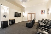 This single-floor conference room is designed to facilitate productive meetings in a well-appointed, light-filled environment. The space is defined by its striking double-height ceilings, which lend an open, airy quality rarely found in standard meeting rooms. A sweeping bank of large windows, dressed with neutral roller shades, lines one wall from floor to ceiling, flooding the interior with abundant natural light and offering views of an outdoor pool area and surrounding palm trees.

The central focus of the room is a substantial oval conference table with a bold red base, surrounded by ergonomic black leather rolling chairs that comfortably seat a full group. A wall-mounted flat-screen television is positioned for easy viewing by all attendees, while a large whiteboard on the adjacent wall supports presentations and collaborative work sessions. Wall sconces provide supplemental lighting, contributing to the room's polished yet functional atmosphere.

A dedicated refreshment station in the corner includes a coffee maker and printer, adding convenience for extended meetings. Entry is provided through a set of glass-paneled double doors, and the carpeted flooring throughout contributes to the room's professional ambiance. Additional seating along the windowed wall accommodates observers or breakout participants.