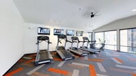 Located at 845 Yard St in Grandview Heights, Ohio, this single-floor fitness center is designed with both functionality and comfort in mind. The space features a clean, contemporary aesthetic with crisp white walls and a vaulted ceiling equipped with track lighting and a ceiling fan for optimal air circulation during workouts. The cardio area is a standout feature, housing a row of treadmills and additional cardio machines, each positioned in front of wall-mounted flat-screen televisions — keeping members entertained throughout their sessions. The flooring is a distinctive geometric-patterned carpet in shades of gray, orange, and blue, adding a bold visual element to the otherwise streamlined space. Large windows along one wall flood the room with natural light while offering views of the surrounding outdoor area, creating an energizing and open atmosphere. Track lighting throughout ensures the space remains well-lit regardless of the time of day. The overall ambiance is modern and motivating, offering a well-appointed environment suited for an effective workout experience.
