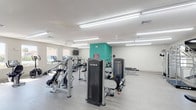 This single-story fitness center offers a spacious, thoughtfully designed layout suited for a full range of workout activities. The main exercise area is generously proportioned, featuring light wood-look flooring throughout and bright white walls accented by a teal feature wall that adds a pop of color to the interior. Overhead, rows of fluorescent strip lighting and ceiling fans ensure the space remains well-lit and comfortable during use.

The cardio zone lines one side of the room, equipped with treadmills, ellipticals, stationary bikes, and a rowing machine, all positioned along a bank of windows that frame outdoor views of palm trees and surrounding greenery, providing an uplifting workout environment. The strength training area is centrally positioned and includes multiple TRUE-brand cable and resistance machines, adjustable benches, and a multi-function cable tower, offering a comprehensive selection for resistance training.

A secondary room provides additional strength equipment, including benches and a cable machine, along with recessed lighting for a focused training environment. Separate open rooms with recessed lighting and wood-look flooring offer flexible space for stretching, group exercise, or other activities.

A kitchenette area with pendant lighting, a stainless steel refrigerator, and wall-mounted water fountains supports the functional needs of users. The overall ambiance is clean, modern, and bright.
