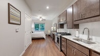 This efficiently designed one-bedroom, one-bathroom unit offers a thoughtful layout that maximizes every square foot of its single-floor footprint. The open living area flows seamlessly from the fully equipped kitchen into the bedroom space, creating a cohesive and functional environment.

The kitchen is a standout feature, outfitted with rich dark-toned shaker-style cabinetry, light granite countertops, a stainless steel gas range with overhead ventilation, a refrigerator, and a sink — all finished in a clean, modern style. White tile backsplash accents the cabinetry wall, adding a bright contrast to the warm cabinetry tones.

Warm hardwood-style flooring runs throughout the entire unit, unifying the space and contributing to its inviting ambiance. The bedroom area is generously proportioned, featuring a ceiling fan with light fixture and a window that frames a lush outdoor tree canopy, allowing natural light to fill the room.

The bathroom maintains the same clean aesthetic with neutral gray walls, wood-style flooring, a shower with curtain, and modern brushed nickel fixtures. Recessed lighting throughout the unit provides consistent, well-distributed illumination. The entry door leads directly into the living and kitchen area, offering a practical and welcoming transition into the space.