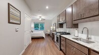 This efficiently designed one-bedroom, one-bathroom unit offers a thoughtful layout that maximizes every square foot of its single-floor footprint. The open living area flows seamlessly from the fully equipped kitchen into the bedroom space, creating a cohesive and functional environment.

The kitchen is a standout feature, outfitted with rich dark-toned shaker-style cabinetry, light granite countertops, a stainless steel gas range with overhead ventilation, a refrigerator, and a sink — all finished in a clean, modern style. White tile backsplash accents the cabinetry wall, adding a bright contrast to the warm cabinetry tones.

Warm hardwood-style flooring runs throughout the entire unit, unifying the space and contributing to its inviting ambiance. The bedroom area is generously proportioned, featuring a ceiling fan with light fixture and a window that frames a lush outdoor tree canopy, allowing natural light to fill the room.

The bathroom maintains the same clean aesthetic with neutral gray walls, wood-style flooring, a shower with curtain, and modern brushed nickel fixtures. Recessed lighting throughout the unit provides consistent, well-distributed illumination. The entry door leads directly into the living and kitchen area, offering a practical and welcoming transition into the space.