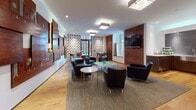 Located at 227 Church St in New Haven, Connecticut, this single-floor space offers a thoughtfully designed communal environment that balances contemporary style with artistic character. The main lounge area is anchored by sleek black leather seating arrangements — including sofas and armchairs — positioned around a glass-topped coffee table atop a textured area rug, with light hardwood flooring extending throughout the surrounding space.

A striking decorative wood panel installation dominates one wall, featuring geometric and symbolic motifs arranged in an eclectic, layered composition that serves as a bold focal point. The opposite end of the lounge transitions into a casual dining or café-style zone, where bar-height tables and modern stools sit beneath bold patterned wallpaper with a graphic floral-tile motif. A richly framed glass door in this area allows natural light to filter in and offers views of a lush outdoor greenspace.

A compact kitchenette is also present, outfitted with warm wood cabinetry, white stone countertops, a stainless steel undermount sink, and a glossy grey subway-tile backsplash. Recessed and flush-mount square ceiling lights provide consistent, even illumination throughout. A wall-mounted flat-screen television adds a functional touch to the lounge. Overall, the space presents a refined yet approachable ambiance.