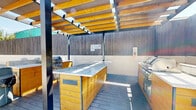 Located at 45 E 12th St in National City, California, this outdoor amenity space is thoughtfully designed to maximize the region's year-round sunshine and mild climate. The grounds are anchored by a spacious pool deck finished with light-toned composite decking, providing a comfortable and stylish surface for lounging. Poolside seating, including Adirondack chairs, invites residents to relax in the open air beneath the Southern California sky, surrounded by mature palm trees that lend a resort-like atmosphere.

Adjacent to the pool area, a generous concrete courtyard serves as an al fresco gathering space, furnished with dining tables and chairs shaded by large umbrellas — ideal for outdoor meals or social gatherings. Lush, manicured hedges and raised planter beds with vibrant foliage line the perimeter, adding greenery and a sense of privacy. Modern concrete block walls and warm wood-panel fencing define the boundaries of the space, contributing to a clean, contemporary aesthetic.

A standout feature is the pergola-covered outdoor kitchen, equipped with a built-in grill and prep station, perfect for barbecues and entertaining. The structure's open-slat roof allows natural light to filter through while providing partial shade. The multi-story building backdrop, with its glass balconies and wood-accented facade, frames the entire outdoor space with a sleek, modern architectural presence.