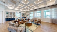 Located at 1730 Savona Point Cir in Cape Coral, Florida, this generously sized single-floor community space offers a well-appointed layout designed for gathering and socializing. The welcoming entryway features soft sage-green walls, patterned carpet flooring, and large glass double doors that flood the space with natural light and provide views of the surrounding grounds.

The main living area is a standout, boasting a striking coffered ceiling with recessed lighting that adds architectural elegance and a sense of grandeur. Light hardwood-style flooring, built-in shelving with navy blue lower cabinetry, and a wall-mounted television create a polished yet comfortable lounge environment. A secondary living area transitions seamlessly from the main space, maintaining the cohesive coastal aesthetic.

The kitchen is a design highlight, featuring bold navy cabinetry with gold hardware, a large quartz-topped island with bar seating, statement globe pendant lights in brushed brass, and stainless steel appliances. Clerestory windows above the cabinetry invite abundant natural light. The adjacent dining area opens directly to an outdoor patio through full-height glass sliding doors and a glass panel door, offering views of palm trees and the outdoor amenity area — perfectly suited to Florida's indoor-outdoor lifestyle.