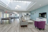 Located at 1410 Hartman Pl in Manhattan, Kansas, this single-floor community clubhouse offers a well-designed, open-concept layout that accommodates a variety of social and recreational activities. The space is unified by light wood-tone flooring throughout, neutral gray and soft teal accent walls, and abundant recessed lighting that contributes to a bright, welcoming atmosphere.

The main gathering area serves as the centerpiece of the space, highlighted by a coffered tray ceiling with a skylight that draws in natural light. This zone features a full-size pool table, a comfortable lounge seating arrangement with a sofa and accent chairs, and ample open floor space that supports flexible use. The area flows seamlessly into a fully equipped kitchen, which includes white shaker-style cabinetry, stainless steel appliances — including a French door refrigerator and microwave — a deep sink, and a large white-topped island with bar-height seating. The kitchen is designed to support group gatherings and casual entertaining.

Adjacent to the main lounge, a dedicated dining and casual seating area features multiple round pedestal tables with modern wood chairs, a plush purple accent chair, and large windows that frame outdoor views and fill the room with natural light. Glass sliding doors connect this area to a separate room, providing flexible, semi-private space. Overall, the clubhouse presents a clean, contemporary aesthetic ideal for community use.