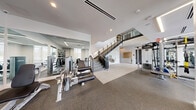 This modern, two-story fitness facility offers a comprehensive workout environment designed for performance and comfort. The main floor — the larger of the two levels — is the heart of the facility, featuring an expansive cardio zone lined with Life Fitness treadmills, ellipticals, and stair climbers positioned along floor-to-ceiling windows that flood the space with natural light and views of the surrounding greenery. Full-length mirrors along the walls visually expand the already generous footprint and allow users to monitor their form throughout their workouts. A dedicated strength training area offers a wide array of cable machines, free weights, benches, and multi-functional rigs, all set on durable rubberized flooring. A sleek, dark-railed staircase connects the main floor to the upper level, where a glass-railed mezzanine overlooks the floor below. The upper level houses an enclosed studio space with light wood flooring, large display screens, and spin bikes, ideal for group fitness or private training sessions. The design throughout is clean and contemporary, with recessed linear lighting, white walls, and an open layout that promotes airflow and visibility across both levels. Practical amenities such as a water fountain station are thoughtfully integrated into the space.