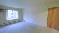 This single-floor space offers a clean, straightforward layout designed for comfortable everyday living. The interior is finished with wall-to-wall carpeting in a neutral tone, providing a soft underfoot feel that extends throughout the space. The walls are painted in a crisp, light neutral shade, creating a bright and open atmosphere that makes the rooms feel airy and welcoming.

Natural light plays a key role in the ambiance of this space. A large window allows an abundance of daylight to filter in, illuminating the interior and offering a view of the surrounding outdoor greenery. The window placement enhances the sense of openness within the room.

Climate comfort is addressed through a baseboard heating system that runs along the lower perimeter of the walls, providing consistent and efficient warmth throughout the colder months. The smooth, white ceiling completes the clean aesthetic of the interior.

Overall, this space presents a simple yet functional environment with a neutral palette that serves as a versatile canvas, allowing for a wide range of interior design and furnishing possibilities. Its straightforward design, natural light, and practical heating system make it a solid and adaptable living space.