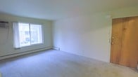 This single-floor space offers a clean, straightforward layout designed for comfortable everyday living. The interior is finished with wall-to-wall carpeting in a neutral tone, providing a soft underfoot feel that extends throughout the space. The walls are painted in a crisp, light neutral shade, creating a bright and open atmosphere that makes the rooms feel airy and welcoming.

Natural light plays a key role in the ambiance of this space. A large window allows an abundance of daylight to filter in, illuminating the interior and offering a view of the surrounding outdoor greenery. The window placement enhances the sense of openness within the room.

Climate comfort is addressed through a baseboard heating system that runs along the lower perimeter of the walls, providing consistent and efficient warmth throughout the colder months. The smooth, white ceiling completes the clean aesthetic of the interior.

Overall, this space presents a simple yet functional environment with a neutral palette that serves as a versatile canvas, allowing for a wide range of interior design and furnishing possibilities. Its straightforward design, natural light, and practical heating system make it a solid and adaptable living space.