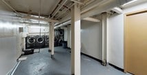 This single-floor utility space presents a practical, no-frills layout typical of a building's lower level. The open floor plan is defined by a series of structural columns that run throughout, supporting an exposed ceiling system of pipes, conduits, and mechanical infrastructure overhead. The painted concrete floors show signs of wear consistent with regular use.

The laundry area is a notable feature of this space, equipped with multiple front-loading washers and stackable dryer units arranged along the walls. The machines are industrial in scale, suggesting shared or communal use. A waste bin is conveniently positioned nearby.

Several solid-core doors are present throughout the space, including at least one marked with a "Private Property – Keep Out" sign, indicating restricted access to utility or storage areas. Additional doorways along a corridor lead to further rooms or storage spaces, visible in the background with emergency exit signage illuminated in red.

The overall ambiance is utilitarian, with fluorescent strip lighting providing functional illumination. White-painted walls and columns contrast with the grey painted floor. The space is clearly designed for operational building functions rather than occupant comfort, prioritizing accessibility and practicality.