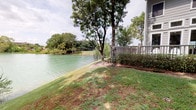 This single-story office space is situated in the Neighborhood Lake neighborhood, offering a serene waterfront setting that sets it apart from a typical workplace environment. The exterior grounds feature a gently sloping lawn that leads directly to the lake's edge, lined with mature trees that provide shade and a tranquil, park-like atmosphere. A wooden deck or fencing structure connects the building to the outdoor surroundings, framing picturesque water views.

Inside, the office is thoughtfully laid out to support a productive work environment. The main office area features vaulted ceilings that lend a sense of openness and airiness, complemented by multiple windows dressed with blinds that allow for controlled natural light. A substantial executive desk with a leather chair serves as the focal point of this workspace, making it well-suited for professional meetings and daily operations.

A dedicated work or copy room houses office equipment, including a commercial-grade multifunction printer, and features teal accent walls that add a pop of color against the neutral-toned interiors. Textured carpet flooring runs throughout the interior spaces, adding warmth underfoot. Tall, lush indoor tropical plants add a refreshing natural element to the interior décor, complementing the scenic outdoor lake views visible from the property.