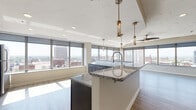 Located at 217 Second St NW in the heart of downtown Canton, Ohio, this contemporary single-floor unit offers a refined urban living experience with high-end finishes and sweeping city views. The open-concept layout seamlessly connects the living and kitchen areas, where a large center island with striking granite countertops and a designer pendant-lit overhead takes center stage. The kitchen is fully equipped with stainless steel appliances — including a refrigerator, range, microwave, and dishwasher — complemented by dark cabinetry, white upper cabinets, and a classic subway tile backsplash. Durable wood-look flooring runs throughout the main living areas, adding warmth to the modern aesthetic. Floor-to-ceiling windows wrap around the space, flooding every room with natural light and offering panoramic views of the Canton skyline. The bedroom features plush carpeting, a ceiling fan, and its own expansive window wall showcasing the cityscape on multiple sides. The bathroom is appointed with granite countertops, a vessel sink, dark wood vanity, large mirror, and a sleek glass-enclosed walk-in shower. Throughout the unit, thoughtful design details — from decorative ceiling textures to contemporary door hardware — create a polished, move-in-ready living environment.
