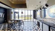 This single-floor space makes a striking first impression with its bold blue-and-white geometric patterned tile flooring, which anchors the entire room with a sense of modern sophistication. Expansive floor-to-ceiling windows line the exterior wall, flooding the interior with abundant natural light and offering sweeping views of the surrounding landscape and greenery beyond. The open layout is thoughtfully arranged to accommodate both dining and social interaction, featuring a large communal table paired with contemporary bar-height chairs — ideal for group gatherings or casual meetings. Overhead, a combination of warm-toned metallic pendant lights and track lighting provides layered illumination, complementing the contrast between the dark ceiling tray and bright white walls. Along one side of the space, a well-appointed kitchenette provides functionality without sacrificing style, complete with a mosaic tile backsplash, sleek cabinetry, a stainless steel sink, and modern appliances. Artwork adorns the walls, adding a curated, gallery-like quality to the space. The overall ambiance is dynamic yet refined, blending vibrant design elements with practical amenities to create an engaging, multipurpose environment.