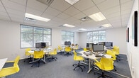 Located in New Orleans, this single-floor business center offers a well-designed, open-plan workspace that balances productivity and comfort. The layout features multiple clusters of white workstations, each equipped with desktop computers and accompanied by ergonomic yellow chairs that add a bold, energetic accent to the otherwise neutral interior palette.

The space is finished with a drop tile ceiling fitted with recessed fluorescent panel lighting, ensuring consistent, even illumination throughout. Gray carpet flooring runs across the entire floor, complementing the clean white walls and contributing to a professional yet inviting atmosphere.

Natural light is a standout feature, with large multi-pane windows fitted with horizontal blinds lining multiple walls. These windows frame views of surrounding greenery, connecting the interior to the outdoors and enhancing the overall brightness of the workspace.

Casual lounge seating—including low-profile sofas with decorative pillows—is integrated into corners of the space, providing informal areas for collaboration or relaxation alongside the primary workstations. Additional dedicated workstations along the perimeter accommodate printers and supplementary equipment.

Colorful artwork punctuates the walls, and the overall design reflects a contemporary, flexible workspace suited for both focused individual work and collaborative activity.