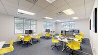 Located in New Orleans, this single-floor business center offers a well-designed, open-plan workspace that balances productivity and comfort. The layout features multiple clusters of white workstations, each equipped with desktop computers and accompanied by ergonomic yellow chairs that add a bold, energetic accent to the otherwise neutral interior palette.

The space is finished with a drop tile ceiling fitted with recessed fluorescent panel lighting, ensuring consistent, even illumination throughout. Gray carpet flooring runs across the entire floor, complementing the clean white walls and contributing to a professional yet inviting atmosphere.

Natural light is a standout feature, with large multi-pane windows fitted with horizontal blinds lining multiple walls. These windows frame views of surrounding greenery, connecting the interior to the outdoors and enhancing the overall brightness of the workspace.

Casual lounge seating—including low-profile sofas with decorative pillows—is integrated into corners of the space, providing informal areas for collaboration or relaxation alongside the primary workstations. Additional dedicated workstations along the perimeter accommodate printers and supplementary equipment.

Colorful artwork punctuates the walls, and the overall design reflects a contemporary, flexible workspace suited for both focused individual work and collaborative activity.