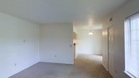 This single-floor residence offers a straightforward, functional layout designed for comfortable everyday living. The living area is finished with neutral wall-to-wall carpeting that extends throughout, contributing to a quiet and cohesive atmosphere. Clean white walls and standard-height ceilings keep the space feeling bright and open, with a simple, uncluttered aesthetic throughout.

The living area flows naturally into a dining space, which is distinguished by hard flooring and illuminated by a classic dome pendant light suspended from the ceiling — a subtle focal point that defines the dining zone. A partial wall separates the living and dining areas, adding a sense of structure without fully closing off the open-plan feel.

Adjacent to the dining area, the kitchen is visible and features wood-toned upper cabinetry and a countertop workspace, providing practical utility in a compact footprint. The transition between the carpeted and hard-floored zones marks a clear delineation between living and functional spaces.

Overall, this residence presents a clean, move-in-ready environment with a neutral palette that offers a versatile canvas for personalization. The open connection between rooms supports an easy flow, making the space feel welcoming and efficiently organized on a single level.