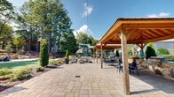 Located at 1113 Powers Ferry Pl SE in Marietta, Georgia, this thoughtfully designed outdoor amenity space offers a resort-like experience surrounded by mature trees and lush, manicured landscaping. The grounds are anchored by two striking wood-framed pavilions with pitched roofs, providing shaded seating areas outfitted with bar-height tables and chairs — ideal for social gatherings. Overhead string lights add a welcoming ambiance that extends into the evening hours.

A generous lawn area serves as the centerpiece for recreation, featuring cornhole boards and open green space for casual outdoor activities. Paved concrete and stone paver walkways connect the various zones of the property, guiding visitors seamlessly between the lawn, seating areas, and surrounding amenities. A resort-style swimming pool with lounge chairs is visible through the mature tree canopy, accessible via a landscaped stairway framed by stone retaining walls.

The grounds are richly planted with Italian cypress trees, ornamental shrubs, crepe myrtles, and towering pines that provide both privacy and a natural backdrop. A dedicated dog-friendly area with fencing, waste stations, and mulched terrain is also present. The building exterior features traditional siding with pitched rooflines, complementing the warm, community-oriented atmosphere of this well-appointed outdoor space.