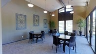 This well-appointed single-floor space functions as an inviting lounge or dining area, thoughtfully designed for comfort and social interaction. The room features impressively high ceilings that create an open, airy atmosphere, further enhanced by a dramatic floor-to-ceiling window wall that floods the interior with natural light and offers sweeping views of the surrounding greenery and landscape.

Several round tables with dark wood finishes are arranged throughout the space, each accompanied by sleek black chairs that lend a modern yet understated elegance. Overhead, a series of elegant bowl-shaped pendant lights hang at varying heights, providing warm, ambient illumination that complements the natural light.

The walls are finished in a soft, earthy green-grey tone, providing a calm and neutral backdrop. Decorative iron-work framed panels with ornate scroll designs add a refined, artistic touch to the walls. A tall potted plant positioned near the window adds a natural, organic element to the décor.

The flooring features a neutral-toned carpet that ties the room together cohesively. Overall, the space strikes a balance between functional and aesthetically pleasing, making it well-suited for casual gatherings, meetings, or dining.