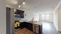 This single-floor residence offers a thoughtfully designed open-concept layout that seamlessly connects the kitchen and living area. The kitchen is a standout feature, boasting dark espresso cabinetry, stainless steel appliances including a refrigerator, range, microwave, and dishwasher, as well as light granite countertops and a peninsula with pendant lighting overhead. Track lighting adds a modern touch, while crown molding along the ceiling lends an elegant finish. Rich hardwood-style flooring runs throughout the main living areas, complementing the neutral wall tones and creating a warm, cohesive aesthetic. The living room is generously proportioned, featuring a ceiling fan, crown molding, a textured ceiling, and large windows that invite abundant natural light. The bedrooms are comfortably sized, finished with plush carpeting, neutral walls, and windows that provide natural light and outdoor views. The bathroom mirrors the kitchen's design language with dark cabinetry, granite countertops, and a large vanity mirror. A dedicated laundry area houses a full-sized washer and dryer with overhead shelving for added storage convenience. Overall, the space presents a clean, modern interior with consistent finishes and a functional, open flow throughout.