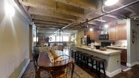 This single-floor loft space showcases a compelling fusion of industrial and rustic design. Exposed brick walls, raw concrete beam ceilings, and visible overhead pipework create a distinctly urban aesthetic that defines the character of the interior throughout.

The open-plan layout seamlessly connects the dining area, living area, and kitchen into one cohesive living space. The dining area is anchored by a warm wooden table surrounded by wrought-iron chairs, set atop a richly patterned area rug that adds texture and warmth to the space. Ambient lighting from a standing floor lamp and track lighting overhead enhances the inviting atmosphere.

The kitchen features dark wood cabinetry, a full suite of appliances including a black refrigerator and range, and a generous breakfast bar with seating for multiple guests. The light-colored countertop provides a clean contrast against the darker elements of the cabinetry and hardware.

Beyond the dining area, the living space is bathed in natural light streaming through large windows, further brightening the otherwise moody, industrial tones of the interior. The combination of exposed structural elements, warm wood tones, brick textures, and thoughtful furnishings gives this loft a welcoming yet architecturally distinctive ambiance.