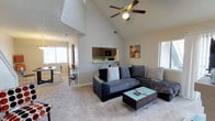 This two-story residence offers two bedrooms and two bathrooms across a generously sized floor plan. The living area immediately impresses with dramatic vaulted ceilings that soar overhead, creating a sense of openness and volume. A ceiling fan is centrally mounted, providing both function and a finishing architectural touch. Plush wall-to-wall carpeting runs throughout the main living space, adding warmth and comfort underfoot.

The open-concept design connects the living room seamlessly to the dining area, which benefits from natural light streaming through multiple windows fitted with blinds. A pass-through window links the living space to the kitchen, where dark wood cabinetry and black appliances are visible, offering a practical and cohesive design aesthetic.

Multiple windows throughout the space ensure consistent natural light, enhancing the airy feel already established by the high ceilings. The two-story layout provides additional flexibility and separation between living and private spaces. Overall, the home offers a comfortable, well-proportioned environment suited for everyday living, with architectural details that elevate the interior beyond the standard.