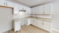 Located at 1150 Vine St in Liverpool, NY, this thoughtfully renovated single-floor space presents a clean, modern aesthetic carried consistently throughout. Light wood luxury vinyl flooring flows seamlessly across every room, lending warmth and continuity to the open layout.

The main living area is generously proportioned, bathed in natural light from a row of wide horizontal windows that frame outdoor views. Recessed ceiling lights and baseboard heating complete the comfortable, functional setup. A secondary room offers similar brightness with its own window and ceiling fixture, making it well-suited as a bedroom or private office.

The kitchen is a standout feature, boasting white shaker-style cabinetry with brushed nickel hardware, granite-look countertops, a subway tile backsplash, and an under-cabinet range hood. The layout makes efficient use of space with ample storage and a stainless steel sink.

The bathroom is elegantly finished with large-format marble-look tile lining the tub surround and accent walls, paired with matte black fixtures that add a contemporary edge. A full soaking tub and overhead shower round out this refined space.

The entry area provides a welcoming transition into the unit, with clean walls, baseboard heating, and easy access to the main living spaces. Overall, this space offers a polished, move-in-ready environment with modern upgrades throughout.