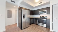 This single-floor unit presents a clean, contemporary design characterized by light wood-tone flooring and bright white walls throughout. The open-concept layout connects the living area to the kitchen seamlessly, creating a sense of spaciousness and ease of movement.

The kitchen is a standout feature, outfitted with a full suite of stainless steel appliances including a side-by-side refrigerator with an in-door ice and water dispenser, a dishwasher, a microwave, and an electric range. Dark espresso-toned cabinetry extends both above and below the granite-patterned countertops, offering ample storage. A louvered closet door adjacent to the kitchen provides additional storage access.

The living area is generously proportioned, bathed in natural light from large windows dressed with vertical blinds. The clean, neutral palette creates a versatile backdrop for any furnishing style.

The bedroom is a calm, light-filled space with wood-look flooring and a window fitted with horizontal blinds that allow for adjustable natural light.

The bathroom features a wide granite-topped vanity with dark cabinetry, sleek hardware, a large wall mirror, and light tile flooring, reinforcing the modern aesthetic carried throughout the unit.