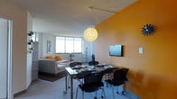 This single-floor space showcases a thoughtfully designed open-plan layout that seamlessly blends the living and dining areas into one cohesive environment. The most striking design element is the bold, warm amber accent wall that anchors the dining zone, creating a vibrant focal point against the crisp white ceiling and neutral flooring. A wall-mounted flat-screen television sits prominently on this accent wall, adding a modern, functional touch.

The dining area features a sleek dark-toned rectangular table surrounded by contemporary black chairs with a streamlined, ergonomic design. A sculptural, globe-shaped pendant light hangs overhead, providing both ambient illumination and a decorative centerpiece that draws the eye upward.

The adjoining living area is furnished with a clean-lined, neutral-toned sofa accented with orange and white throw pillows that echo the warm color palette of the dining space. A striped area rug grounds the seating arrangement and adds textural contrast to the floor. Large windows flood the space with natural light, offering views of the surrounding cityscape and enhancing the open, airy feel of the interior.

A tall, vertical decorative element near the window adds an artistic accent, while framed wall art contributes to the curated, contemporary character of the overall space.