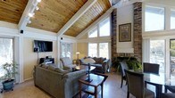 This inviting single-floor space makes a striking first impression with its dramatic vaulted ceilings clad in warm, natural wood planking and framed by crisp white structural beams. The open layout is anchored by a commanding stone fireplace that rises toward the peak of the ceiling, adding a rustic yet refined focal point to the living area. Track lighting mounted along the ceiling beams provides both functional illumination and an architectural accent throughout the space.

The living area is generously proportioned, offering ample room for multiple seating arrangements. Comfortable sofas and accent chairs are thoughtfully arranged to encourage conversation while maintaining clear sightlines to the wall-mounted flat-screen television. Large windows on multiple walls flood the interior with natural light and frame views of the surrounding outdoors, enhancing the open, airy atmosphere.

Light hardwood-style flooring runs throughout, complementing the warm tones of the wood ceiling and stone fireplace. A dining area with a round table and chairs sits adjacent to the living space, creating a natural flow between areas. Decorative touches such as sculptural wall art, potted plants, and layered lighting contribute to the welcoming, cabin-inspired ambiance. Overall, the space balances rustic character with comfortable, everyday livability.