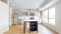 This single-floor space is designed with a clean, modern aesthetic that emphasizes functionality and a neutral color palette throughout. The kitchen serves as the focal point of the layout, outfitted with gray shaker-style cabinetry, white quartz countertops, and a full suite of stainless steel appliances, including a refrigerator, electric range, built-in microwave, and dishwasher. A hexagonal tile backsplash adds subtle texture, while a deep double-basin sink with a sleek gooseneck faucet provides practical utility. A central island with a dark wood base and open shelving offers additional workspace and storage. Large windows in the kitchen area allow natural light to fill the space, creating a bright and airy atmosphere. Light hardwood-style flooring flows throughout the main living and kitchen areas, contributing to the cohesive, open feel of the floor plan. The bedroom features neutral carpet and a sliding closet door with matte black hardware, maintaining the consistent modern style. The full bathroom is finished with the same light flooring, gray shaker vanity cabinetry, a white countertop, and a tub-shower combination with matte black accents on fixtures and hardware, lending a polished, contemporary touch throughout.