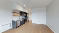 This single-floor unit showcases a contemporary open-plan layout where the kitchen flows seamlessly into the living area. The kitchen is thoughtfully designed with dark cabinetry contrasted against a classic white subway tile backsplash, creating a clean, modern aesthetic. Floating natural wood shelves add a warm, industrial touch above the counter space. Stainless steel appliances, including a refrigerator with ice and water dispenser, microwave, electric range, and dishwasher, equip the kitchen with full functionality.

Light-toned hardwood flooring extends throughout the entire space, lending warmth and visual continuity. The open living area adjacent to the kitchen offers a generous, flexible space with clean white walls and recessed lighting overhead, providing a bright and neutral canvas. The ceilings are notably tall, enhancing the sense of openness throughout the unit. A sliding door visible in the background hints at additional storage or entryway access. Overall, the space presents a minimalist, contemporary ambiance with a balanced mix of natural warmth and modern finishes.