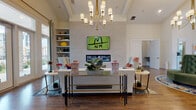 Located at 900 Graces Reserve Circle in Concord, NC, this single-floor community clubhouse combines sophisticated design with a warm, welcoming atmosphere. Soaring ceilings throughout create an expansive sense of openness, while rich hardwood flooring anchors each area with timeless warmth.

The main lounge area serves as the centerpiece of the space, featuring a wall-mounted fireplace flanked by built-in shelving and cabinetry, ideal for displaying décor and books. A statement chandelier with gold accents hangs overhead, complementing the coffered ceiling detail and adding an upscale touch. Comfortable seating arrangements are thoughtfully positioned to encourage conversation and relaxation.

A bright and airy sitting area is bathed in natural light, courtesy of floor-to-ceiling glass doors that open to outdoor views of the surrounding community. The large paned windows and elegant drapery panels enhance the sense of connection between interior and exterior spaces.

Adjacent to the main lounge, a stylishly appointed bar area features a granite countertop, tiled backsplash, and plush navy blue upholstered barstools. Pendant lighting above the counter adds a refined, intimate ambiance, while a beverage station provides added convenience.

The entry foyer welcomes visitors with a sculptural pedestal table, vibrant botanical accents, and direct exterior access through glass-paneled doors. Throughout the clubhouse, curated artwork, layered area rugs, and bold accent pieces contribute to a vibrant yet polished aesthetic.
